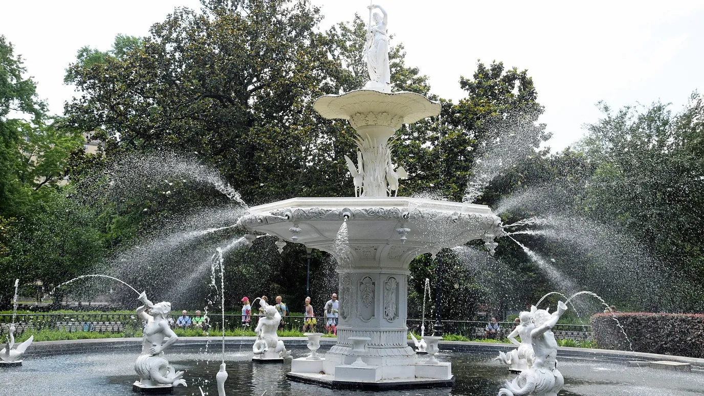 The white Forsyth Park fountain in Savannah, with water jets and oak trees behind it.